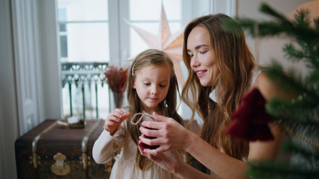 Tender woman giving xmas toy to girl indoors closeup. Smiling mom decorating Christmas tree with daughter at cozy room. Curious little baby putting decorations at green fir branch. New Year conceptの写真素材