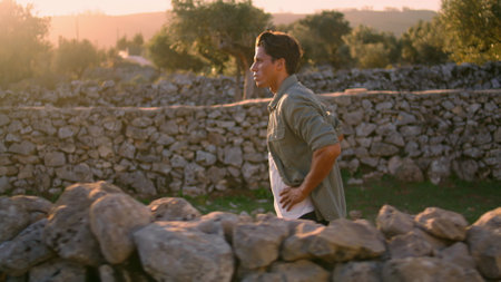 Calm man strolling ancient fence landscape side view. Resting farm worker walking alone enjoying break at countryside. Brunette guy walking at olive trees plantation closeup. Male person at gardenの写真素材