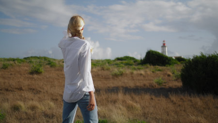 Blonde woman watching lighthouse at meadow closeup. Cute tourist walking in autumn nature landscape. Calm girl stepping field relaxing outdoors. Traveling lady looking distance enjoying vacationの写真素材