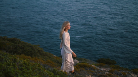 Thoughtful woman walking ocean cliff alone. Dreaming girl stepping marine view hill at summer evening. Relaxed lady rest enjoying beautiful sunset nature. Blonde model travelling sea water landscapeの写真素材