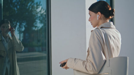 Young business woman taking off wireless earbuds watching window reflection outdoors. Beautiful stylish girl looking camera standing city street sunny day close up. Cute lady face with natural makeupの写真素材