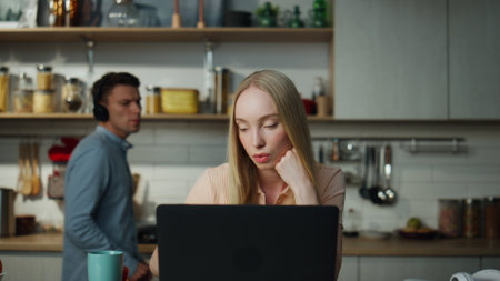 Attractive young businesswoman surfing internet on modern laptop sitting kitchen while smiling husband listening music on backdrop. Focused blond woman developing online business working at home.の写真素材