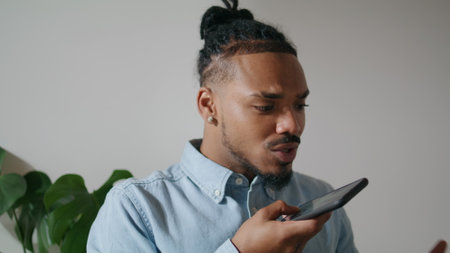 Contemporary man speaking phone at home office closeup. Positive african american freelancer recording smartphone voice message. Black hair guy talking mobile speakerphone working at remote studioの写真素材