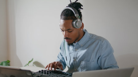 Contemporary arranger creating track apartment closeup. Inspired music producer touching midi keyboard looking laptop. African american headphones man using controller audio equipment working studioの写真素材