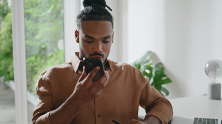 Attentive freelancer listening smartphone voice message indoors closeup. Modern guy using cellphone sitting table alone. African american person sticks to ear loudspeaker looking phone screen at homeの写真素材