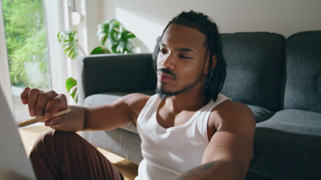 Smiling man sketching paper at apartment portrait. Satisfied brown skin guy holding brush looking masterpiece. African american artist sitting floor zoom out. Painter creating at domestic atmosphereの写真素材