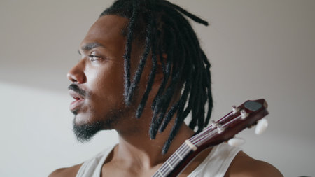 Thoughtful guy enjoying ukulele at light apartment closeup. Involved man improving music skills playing song at home. African american musician holding guitar resting with instrument. Hobby conceptの写真素材