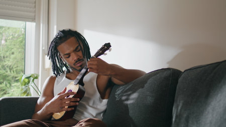 Focused musician playing guitar in domestic place. Contemporary person touching ukulele strings sitting sofa. Satisfied man enjoying music in apartment. African american guy extracting sound at homeの写真素材