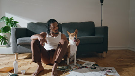 Positive man posing pet at home zoom on. African american guy embracing yawning dog sitting floor portrait. Smiling artist relaxing after drawing closeup. Modern painter resting with domestic animalの写真素材