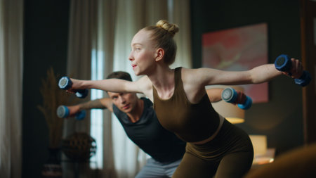 Young family doing gym exercise with dumbbells at home. Sporty attractive couple making hands swing with weight. Slim blond woman training arms strength with athletic man. Partners workout together.の写真素材