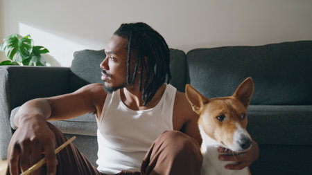 Thoughtful guy embracing dog at sofa room closeup. African american man petting domestic animal sitting wooden floor. Brown skin artist relaxing after drawing sketches at home. Modern painter restingの写真素材