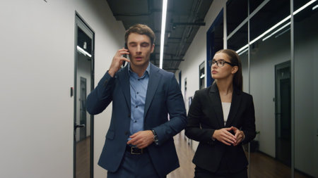 Confident colleagues going corridor in suits. Successful businessman making call discussing project schedule arrangements. Serious management team walking together. Partnership communication conceptの写真素材