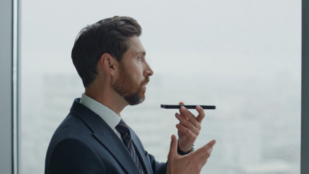 Confident bearded man speaking into speaker phone standing near office window close up. Side view of attractive serious manager have phone conversation using loudspeaker. Smart businessman working.の写真素材