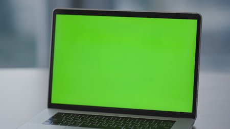 Empty green screen monitor of modern laptop standing on office desk close up. Turned on chroma key...