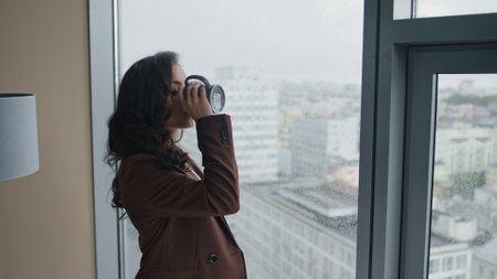 Confident young businesswoman drinking coffee standing in modern office looking at panoramic window close up. Brunette woman thinking about business ideas career advancement. Girl making work break.の写真素材