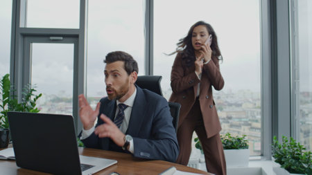 Two businesspeople busy at work in modern luxury office. Emotional businessman reading great news on laptop rejoicing success. Attractive brunette ceo woman talking smartphone solving company issues.の写真素材