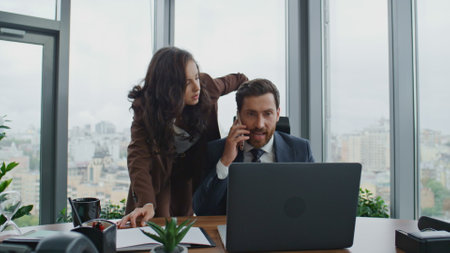 Busy colleagues solving company problems on phone conversation at office. Bearded business man talking telephone while brunette businesswoman listening looking laptop. Smart partners working together.の写真素材
