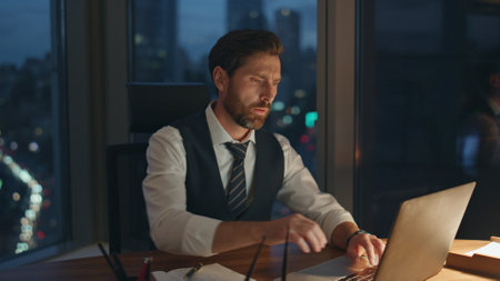 Tired entrepreneur focused on typing laptop working at modern office late night close up. Overworked executive manager feeling headache having overtime. Serious bearded businessman busy of job tasks.の写真素材