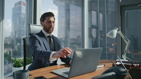 Confident bearded business man finish laptop work shutting notebook lid. Stylish manager getting up from wooden desk taking break. Serious boss looking on panoramic window modern office at skyscraper.の写真素材