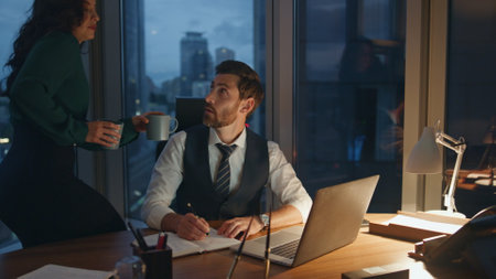 Focused successful coworkers discussing business. Girl assistant bringing drink to overworked businessman sitting at office laptop late evening. Busy manager taking cup coffee from young colleagueの写真素材