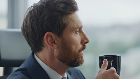 Thoughtful businessman taking break drinking coffee at corporate office close up. Side view of calm confident manager tasting beverage resting from work looking at window cabinet. Business concept.の写真素材