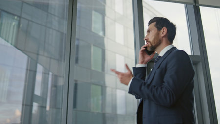 Serious boss talking mobile phone at office wearing stylish suit close up. Stressed businessman having smartphone conversation indoors. Nervous ceo manager speaking gesturing at panoramic windows.の写真素材