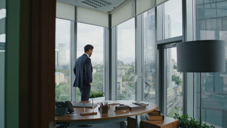 Thoughtful bearded entrepreneur looking at office window standing in modern cabinet. Confident smart businessman thinking business ideas planning future investment strategy. Ceo manager working alone.の写真素材