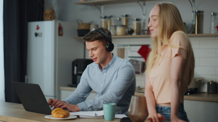 Young music producer working at home kitchen using laptop wearing wireless headphones. Attractive blond wife listening melody enjoying husband work. Cute married couple have fun together in apartment.の写真素材