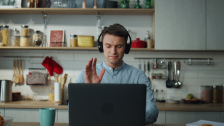 Attractive remote worker saying goodbye to colleagues after video conference close up. Young man in headphones closing laptop drinking coffee sitting at home kitchen. Manager finishing online meeting.の写真素材