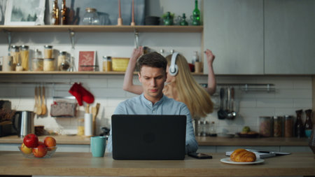 Focused creative writer working home on laptop drinking coffee. Young active woman wearing headphones dancing watching on husband computer. Calm serious man looking on screen ignoring cheerful wife.の写真素材