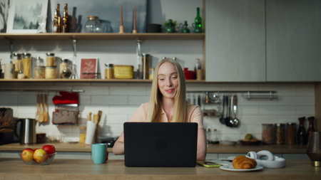 Relaxed attractive woman finish video call closing laptop on kitchen table. Young businesswoman saying goodbye waving hand in computer web camera. Girl manager working remotely at apartment.の写真素材