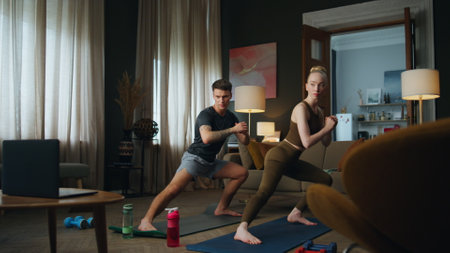 Fit man workouting with slim woman in room watching video lesson. Sporty family couple training body on mat at home. Young guy exercising with blond girl stretching legs. Fitness practice togetherの写真素材