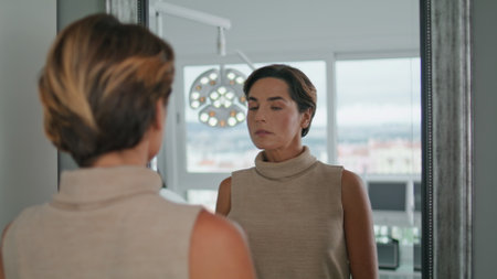 Middle-aged woman looking mirror standing in modern cosmetology clinic close up. Attractive elegant lady checking results anti-aging aesthetic procedures in beauty salon. Client examining face skin.の写真素材