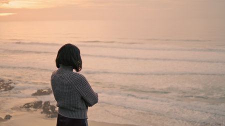Lonely melancholy girl looking on cold gray ocean waves standing alone at seashore autumn evening. Sad attractive brunette girl posing on empty beach wearing sweater. Loneliness tranquility concept.の写真素材