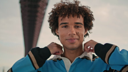 Smiling teenager posing camera at street fashion portrait. Positive zoomer standing looking alone closeup. Curly hair guy demonstrating emotions at city settings. Casual student face staring lensの写真素材
