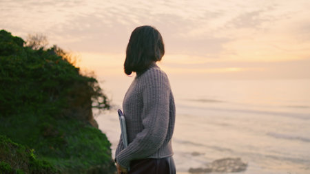 Dreamy pensive woman enjoy ocean twilight walking on coast hill holding album. Attractive thoughtful girl posing in front beautiful sunset sky. Young meditative brunette relax on gloomy nature alone.の写真素材