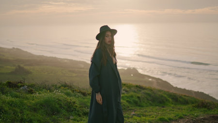 Fashion young woman posing at beautiful sunset wearing stylish hat. Attractive serious model standing on hill green grass near calm ocean. Confident elegant girl looking camera taking off dark coat.の写真素材