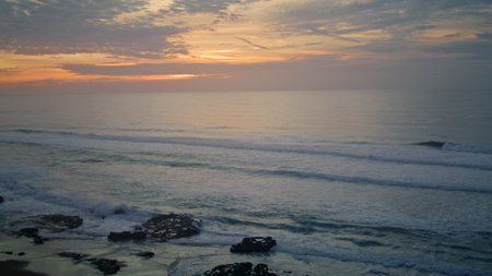 Empty moody evening beach view at sunset. Calm ocean waves washing sandy coast with stones. Picturesque view of cloudy sky with orange sundown over gray sea. Overcast twilight weather on seashore.の写真素材