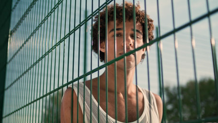 Young sportsman posing basketball court portrait. Curly bad guy looking camera with ball in hands alone. Serious teenager face staring lens on sport ground. Young man leaning stadium grid at summerの写真素材