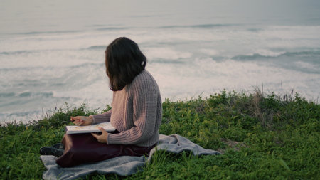 Lonely brunette sitting on blanket with book looking gloomy seascape alone. Attractive young woman relax on nature enjoying calm evening nature. Dreamy romantic girl feeling harmony tranquility.の写真素材