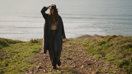 Relaxed stylish girl walking green stony coast at sunny autumn evening. Young woman wearing elegant coat going on grass holding hat from windy weather. Happy brunette enjoying beautiful view oceanの写真素材
