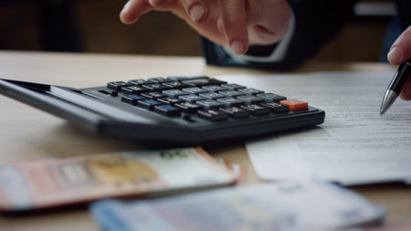 Unknown man using calculator counting corporate budget indoors close up. Male hands pressing calculator buttons analyzing business income. Accountant working at table with documents bills money cash.の写真素材