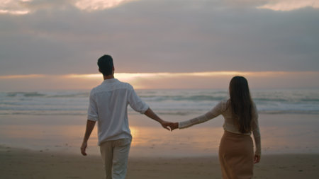 Young spouses relaxing sunbeams beach at cloudy evening. Happy lovers holding hands walking together at sea coast back view. Loving woman man romantic honeymoon date at ocean side. Love relationshipの写真素材