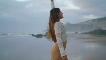 Romantic woman walking evening seashore. Positive lady raising hands at summer ocean vacation. Female person enjoying freedom at marine nature. Dark clouds covered stormy sky. Travelling conceptの写真素材