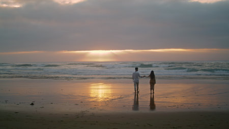 Affectionate newlyweds going sunrise sea shore. Loving woman holding man hand on morning sky beach. Married people watching ocean horizon back view. Love sweethearts enjoying romantic time togetherの写真素材