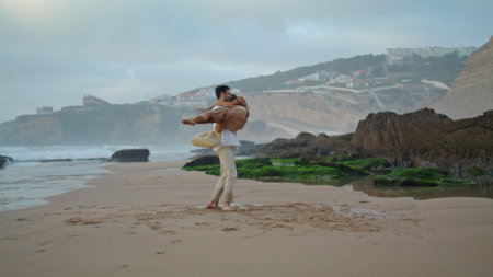 Love couple having fun in marine vacation. Gentle man carrying wife in arms on sea beach. Affectionate hugging couple swirling under cloudy sky. Cheerful lovers celebrate honeymoon together wide shotの写真素材