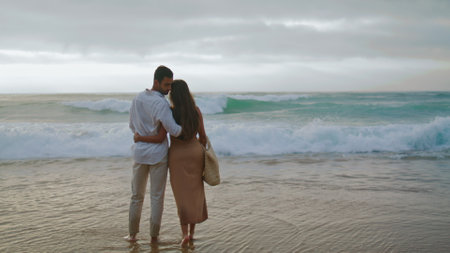Tender family looking sunrise sea beach. Unrecognizable people enjoy time at morning shoreline. Back view cuddling couple touching together at ocean vacation. Peaceful partners rest at calm waterの写真素材