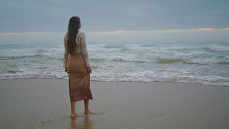 Romantic girl looking ocean view at evening. Dreamy woman walking on stormy beach wide shot. Female model spending vacation at seaside alone. Brunette lady enjoying of scenery marine landscapeの写真素材