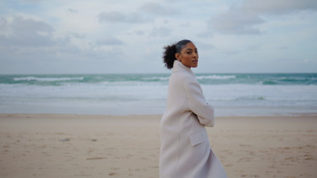 Lonely girl posing ocean beach waves. Serious black hair woman enjoy solitude on autumn weekend. Peaceful african american taking break make decision at quiet place. Serene traveler rest sea shore.の写真素材