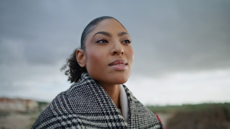 Closeup calm woman walking autumn beach. Black hair attractive girl rest alone looking camera at cloudy sky. Serene african american enjoying break relaxing outdoors. Peaceful female strolling weekendの写真素材